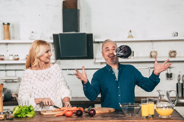 Cheerful woman cutting carrot and looking at red cabbage in air near husband — Stock Photo