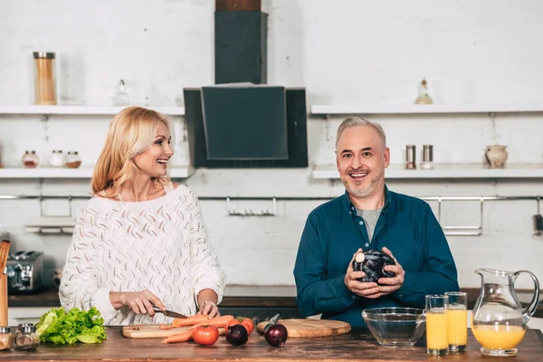 Mujer alegre cortando zanahoria y mirando marido feliz sosteniendo col roja - foto de stock