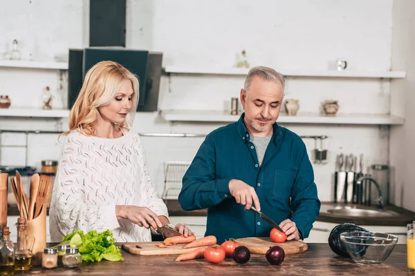 Mulher atraente cortando cenoura perto do marido segurando faca perto de tomate na cozinha — Fotografia de Stock