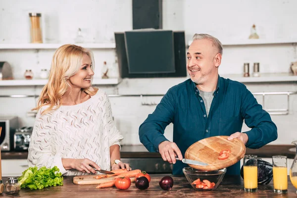 Fröhliche Frau, die Möhren schneidet und glücklichen Ehemann ansieht, der geschnittene Tomaten in Glasschale legt — Stockfoto