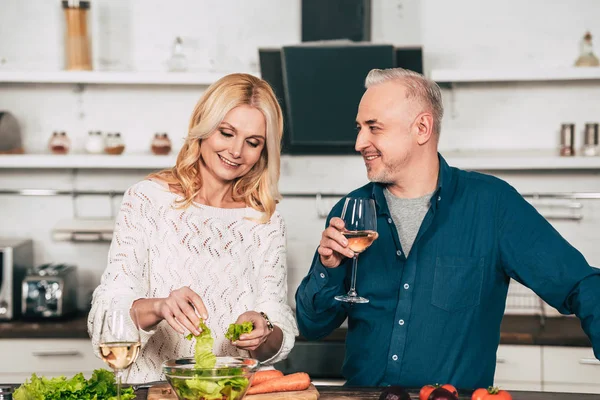 Hombre guapo sosteniendo vaso con vino y mirando a la atractiva esposa rubia sosteniendo hojas de lechuga verde - foto de stock