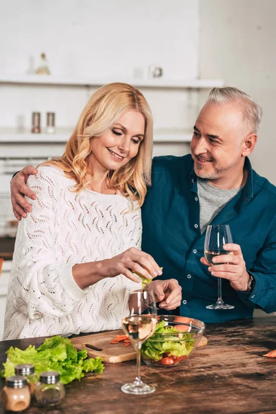Красивый мужчина с бокалом вина обнимает привлекательную блондинку жену держа зеленые листья салата — стоковое фото