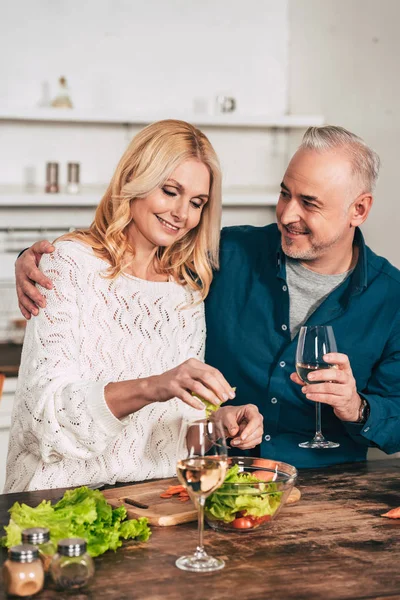 Hombre guapo con copa de vino abrazando atractiva esposa rubia sosteniendo hojas de lechuga verde — Stock Photo