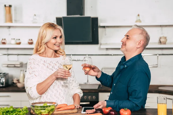 Cheerful couple holding glasses of wine near vegetables in kitchen — Stock Photo