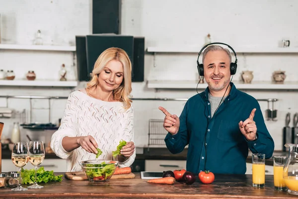 Весела жінка готує їжу біля щасливого чоловіка, слухаючи музику в навушниках на кухні — стокове фото