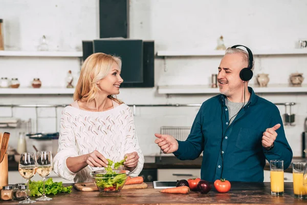 Glückliche Frau bereitet Essen in der Nähe glücklicher Ehemann hört Musik in Kopfhörer in der Küche — Stockfoto