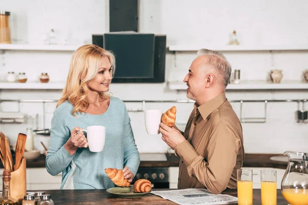 Uomo allegro che tiene gustoso croissant e tazza di bevanda mentre guarda moglie felice — Foto stock