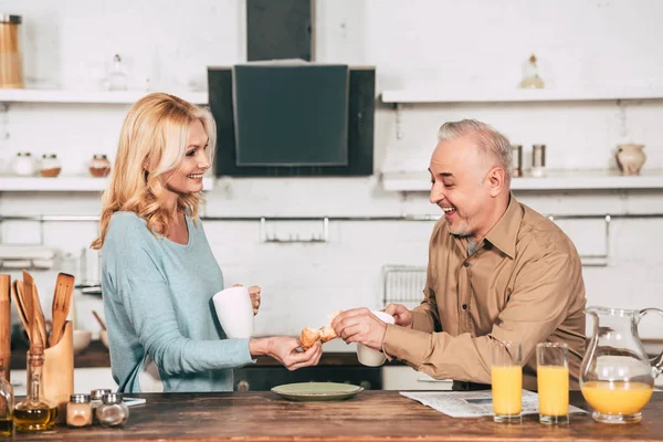 Веселая женщина, делящая вкусный круассан с мужем, держа чашку напитка — стоковое фото