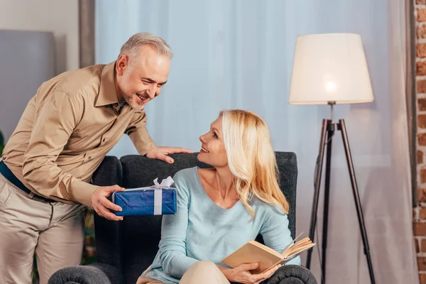 Attractive woman sitting with book and smiling near husband holding gift box in hand — Stock Photo