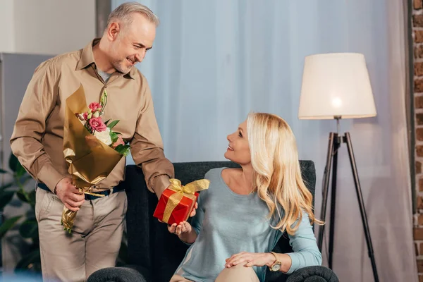 Mujer feliz sentada con caja de regalo y sonriendo cerca de marido alegre sosteniendo flores - foto de stock