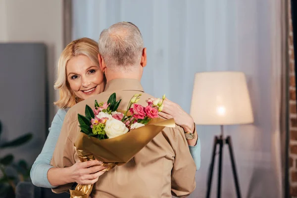 Femme gaie souriant tout en embrassant mari et tenant des fleurs et boîte cadeau dans les mains — Photo de stock