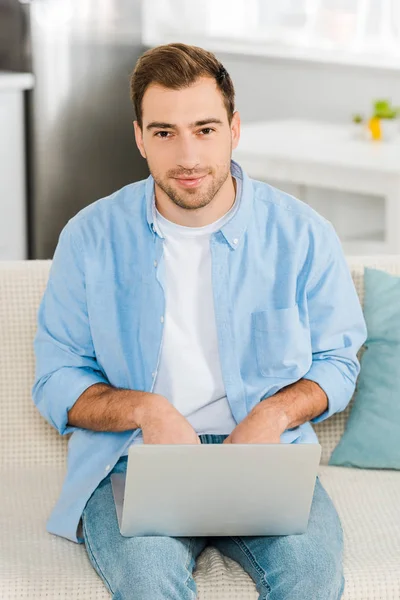 Hombre guapo en ropa casual sentado, mirando a la cámara y utilizando el ordenador portátil en casa - foto de stock