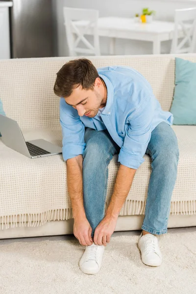 Мужчина сидит на диване с ноутбуком и завязывает шнурки на кроссовках дома — стоковое фото