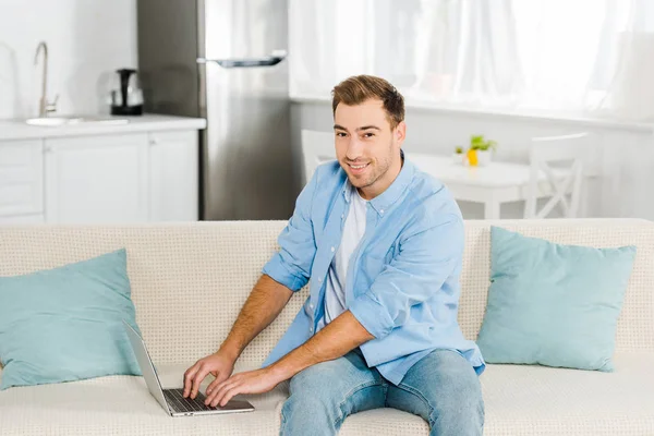 Красивый мужчина, сидящий на диване, смотрящий в камеру и использующий ноутбук дома — стоковое фото