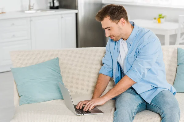 Homem bonito sentado no sofá e usando laptop em casa — Fotografia de Stock