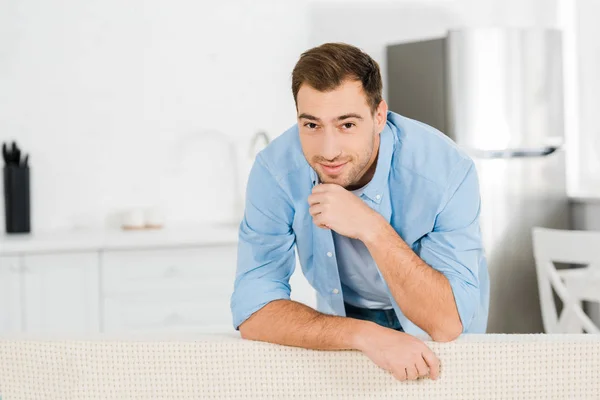 Handsome smiling man propping chin with hand, leaning on sofa and looking at camera at home — Stock Photo