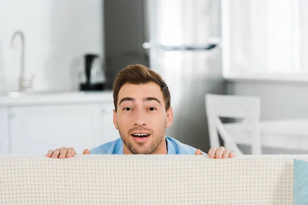 Überrascht schöner Mann schaut auf Kamera und versteckt sich hinter Couch zu Hause — Stockfoto