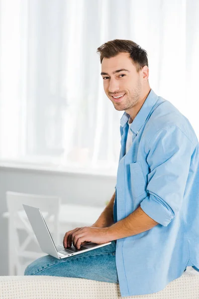 Bel homme souriant assis, regardant la caméra et utilisant un ordinateur portable à la maison avec de l'espace de copie — Photo de stock