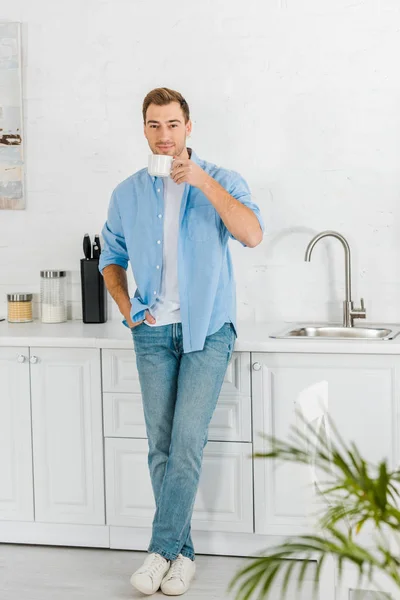 Bell'uomo in abiti casual guardando la macchina fotografica e bevendo caffè in cucina — Foto stock