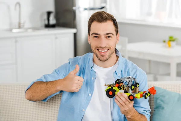 Hombre sonriente guapo mirando a la cámara, sosteniendo el coche de juguete y mostrando el pulgar hacia arriba signo en casa - foto de stock