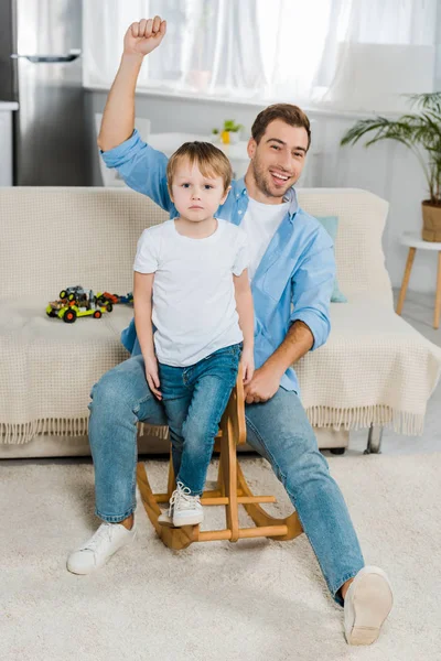 Pai alegre gesticulando com a mão e montando cavalo de balanço de madeira com filho pré-escolar em casa — Fotografia de Stock
