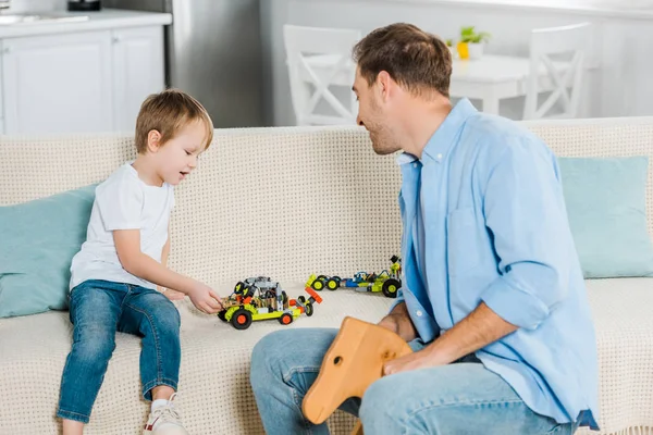 Father riding wooden rocking horse while preschooler son playing with toy cars at home — Stock Photo