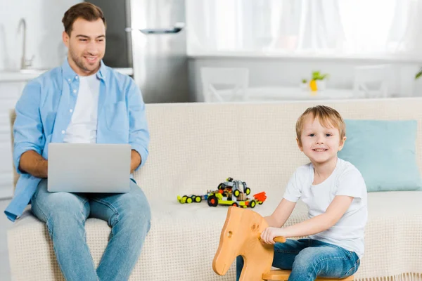 Happy preschooler son riding wooden rocking horse while father using laptop on couch at home — Stock Photo
