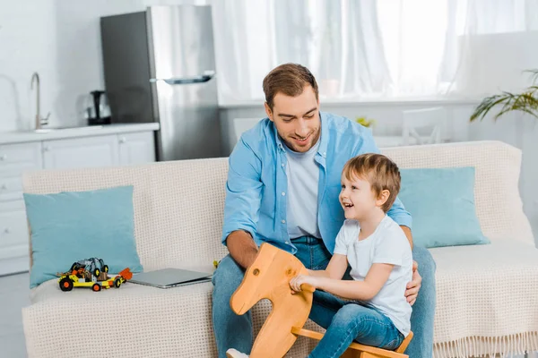 Felice bambino in età prescolare cavalcando cavallo a dondolo in legno mentre sorridente padre seduto sul divano a casa — Foto stock