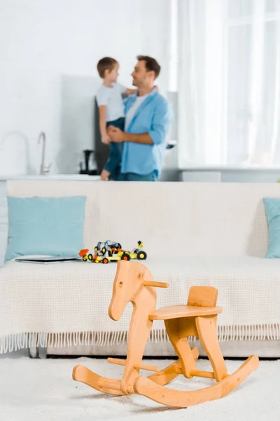 Selective focus of wooden rocking horse, couch with toy cars and father holding son on background at home — Stock Photo