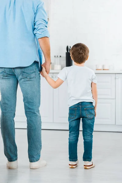 Vista posterior de preescolar cogido de la mano con el padre en la cocina - foto de stock