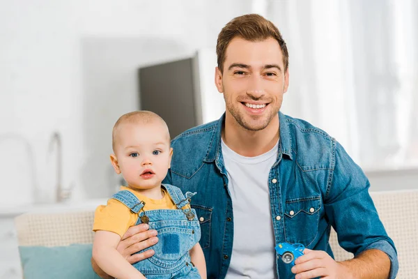 Happy father looking at camera and sitting with adorable toddler son on couch at home — Stock Photo