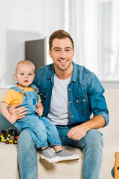 Glücklicher Vater blickt in die Kamera und sitzt mit entzückendem Kleinkind-Sohn auf der heimischen Couch — Stockfoto