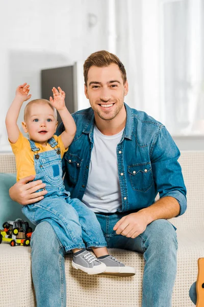 Felice padre seduto sul divano e abbracciare adorabile figlio bambino con le mani in aria a casa — Foto stock