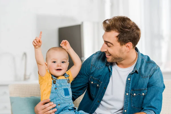 Happy father sitting on couch and hugging adorable toddler son with hands in air at home — Stock Photo