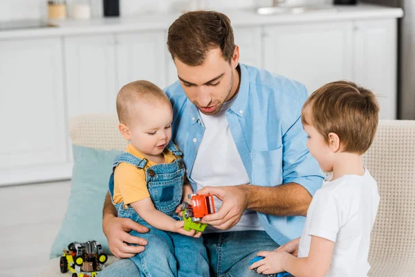 Schöner Vater sitzt auf der Couch, während süße Vorschulkinder und Kleinkinder zu Hause mit Spielzeugautos spielen — Stockfoto