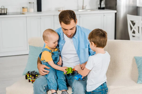Bonito pai sentado no sofá enquanto pré-escolar e crianças filhos brincando com carros de brinquedo em casa — Fotografia de Stock