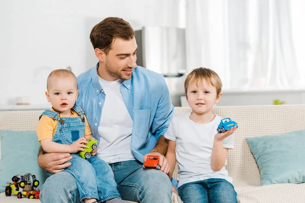 Glücklicher Vater, der auf der Couch sitzt, während Vorschulkinder und Kleinkinder Spielzeugautos zu Hause halten — Stockfoto