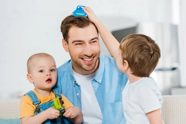 Niños en edad preescolar y niños pequeños sosteniendo coches de juguete y jugando con el padre feliz en casa — Stock Photo