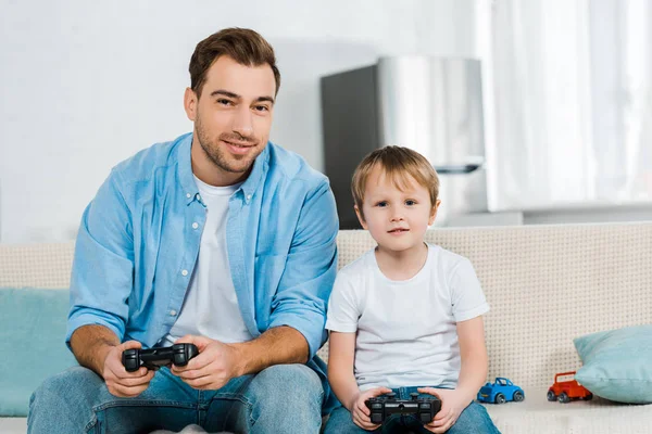 Padre e hijo preescolar sentado con joysticks, mirando a la cámara y jugando videojuegos en casa - foto de stock