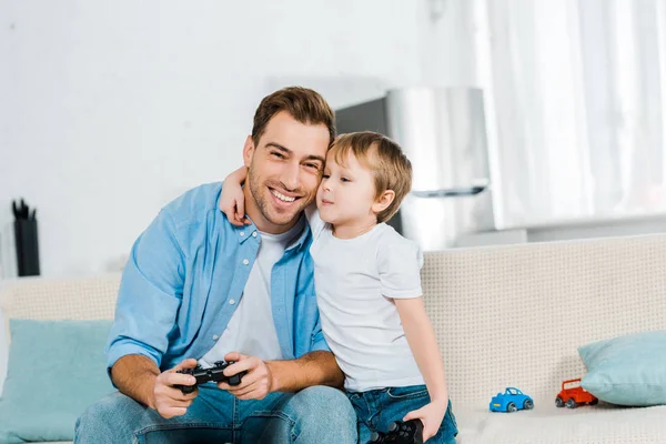 Дошкольный сын с джойстиком обнимает улыбающегося отца во время видеоигры дома — стоковое фото