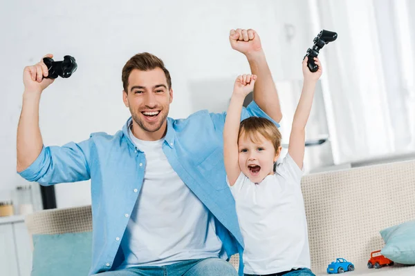 Excited father and preschooler son cheering with hands in air while playing video game at home — Stock Photo