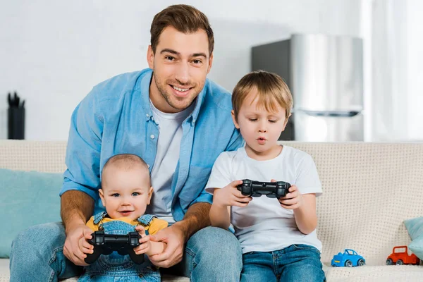Улыбающийся отец играет в видеоигры с очаровательным дошкольником и детьми, сидя дома на диване — стоковое фото