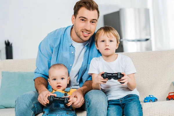 Smiling father playing video game with preschooler and toddler sons while sitting on couch at home — Stock Photo