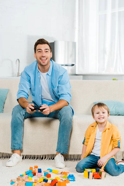 Красивый улыбающийся отец играет в видеоигру, в то время как милый дошкольник сын играет с красочными строительными блоками дома — стоковое фото
