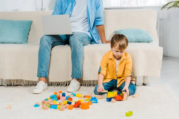 Отец сидит на диване и с помощью ноутбука в то время как дошкольник сын играет с красочными блоками здания дома — стоковое фото