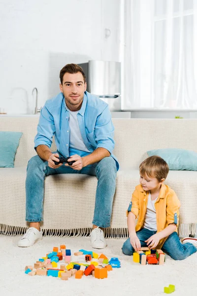 Schöner Vater mit Steuerknüppel spielt Videospiel, während süßer Vorschulsohn zu Hause mit bunten Bausteinen spielt — Stockfoto