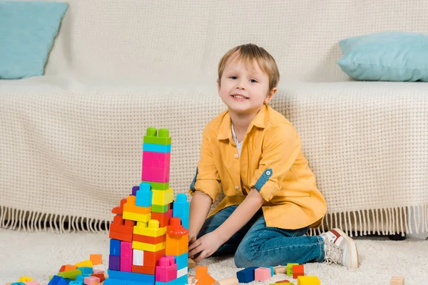 Adorable niño preescolar mirando a la cámara mientras juega con bloques de construcción de colores en casa - foto de stock