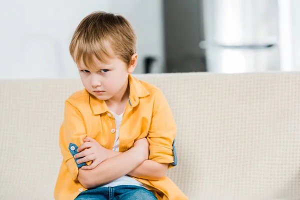 Недовольный дошкольник сидит со скрещенными руками и смотрит в камеру дома — стоковое фото