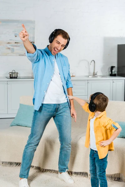 Feliz padre con hijo preescolar en auriculares escuchando música y señalando con los dedos en casa - foto de stock