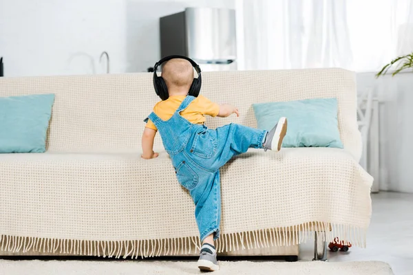 Back view of male toddler in headphones listening music and climbing on couch at home — Stock Photo
